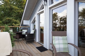 An outdoor deck with wicker furniture, a table with glasses and a bottle, and a view of the surrounding trees.