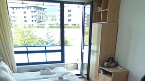 A hotel room with a bed, a desk, a chair, a small table, a cabinet, and a view of buildings outside. - 1 Bed - Sleeps 4 - Pets - Mountain Views (Razlog)