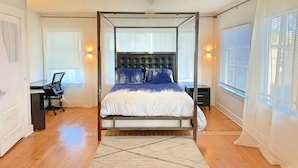 A four-poster bed with a tufted headboard, a desk with a chair, and a large window with blinds. - Heavenly Beach-front Apartment in Venice (Los Angeles)