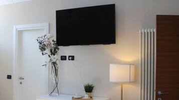 Quarto com cômoda branca, televisão de tela plana na parede, pequena mesa com uma planta e um abajur, e um radiador.