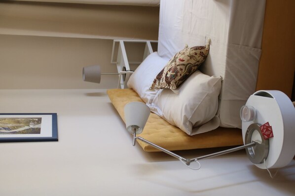 Una cama bien hecha con una almohada, una lámpara de noche y una mesita de noche blanca con un dispensador de pañuelos.