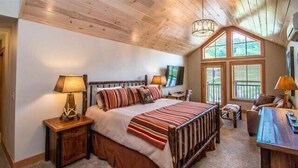 A cozy bedroom with a large bed, two bedside tables, a desk, and a window with blinds.