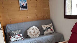 A cozy room with a wooden wall, a grey sofa with skiing-themed pillows, and two wooden chairs with red cushions. - Appartement F2 Avec Large Balcon (Embrun)