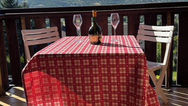 A table with a red and white checkered tablecloth, two wine glasses, and a bottle of wine on a balcony with a mountain view. - Appartement F2 Avec Large Balcon (Embrun)