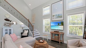 A spacious living room with a staircase, a flat-screen TV, and a large window with blinds. - Bethany Cottage (Port St. Joe)