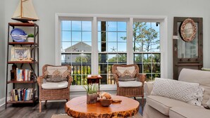 A living room with two wicker chairs, a round coffee table, a bookshelf, and a large window with a view. - Bethany Cottage (Port St. Joe)