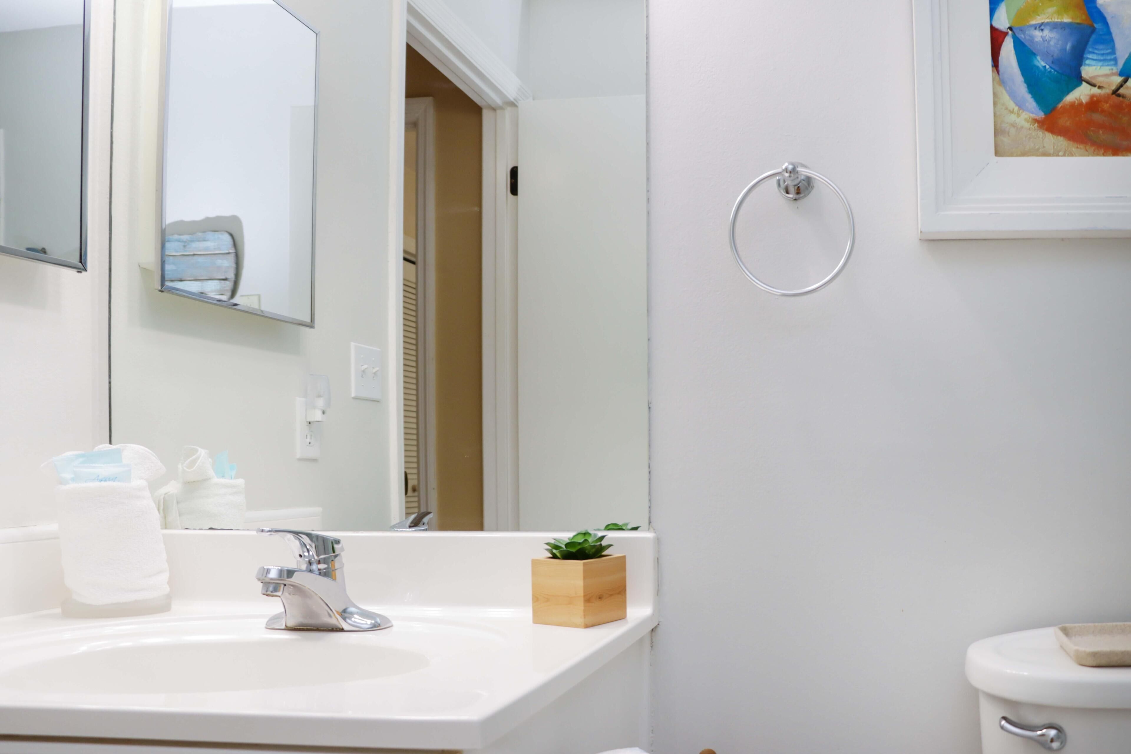 Une salle de bain avec un lavabo, un miroir et des toilettes, le tout de couleur blanche. On trouve une petite boîte en bois sur le lavabo et un porte-serviettes au mur.