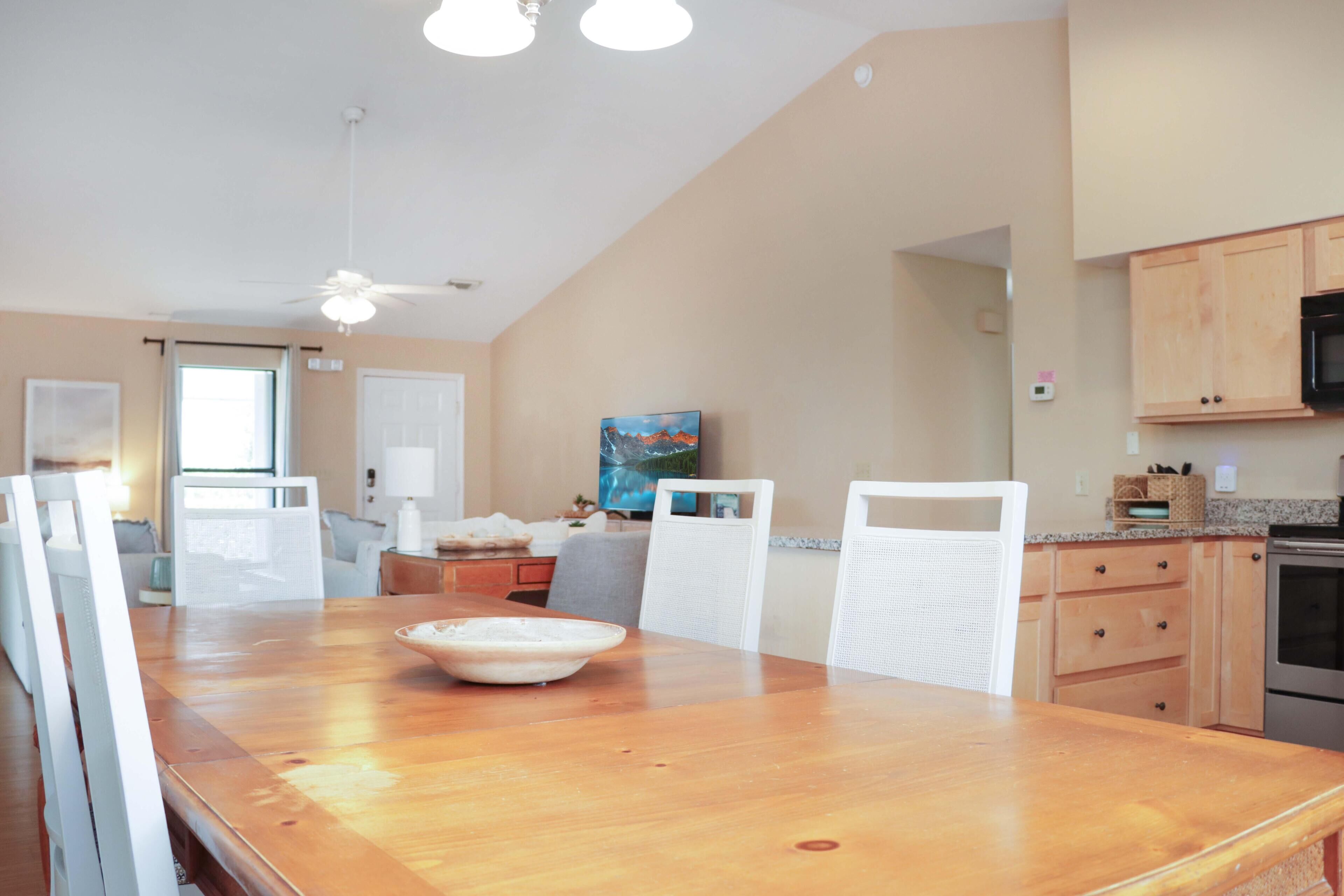 A dining area with a wooden table, white chairs, a ceiling fan, and a kitchen with wooden cabinets and a microwave.