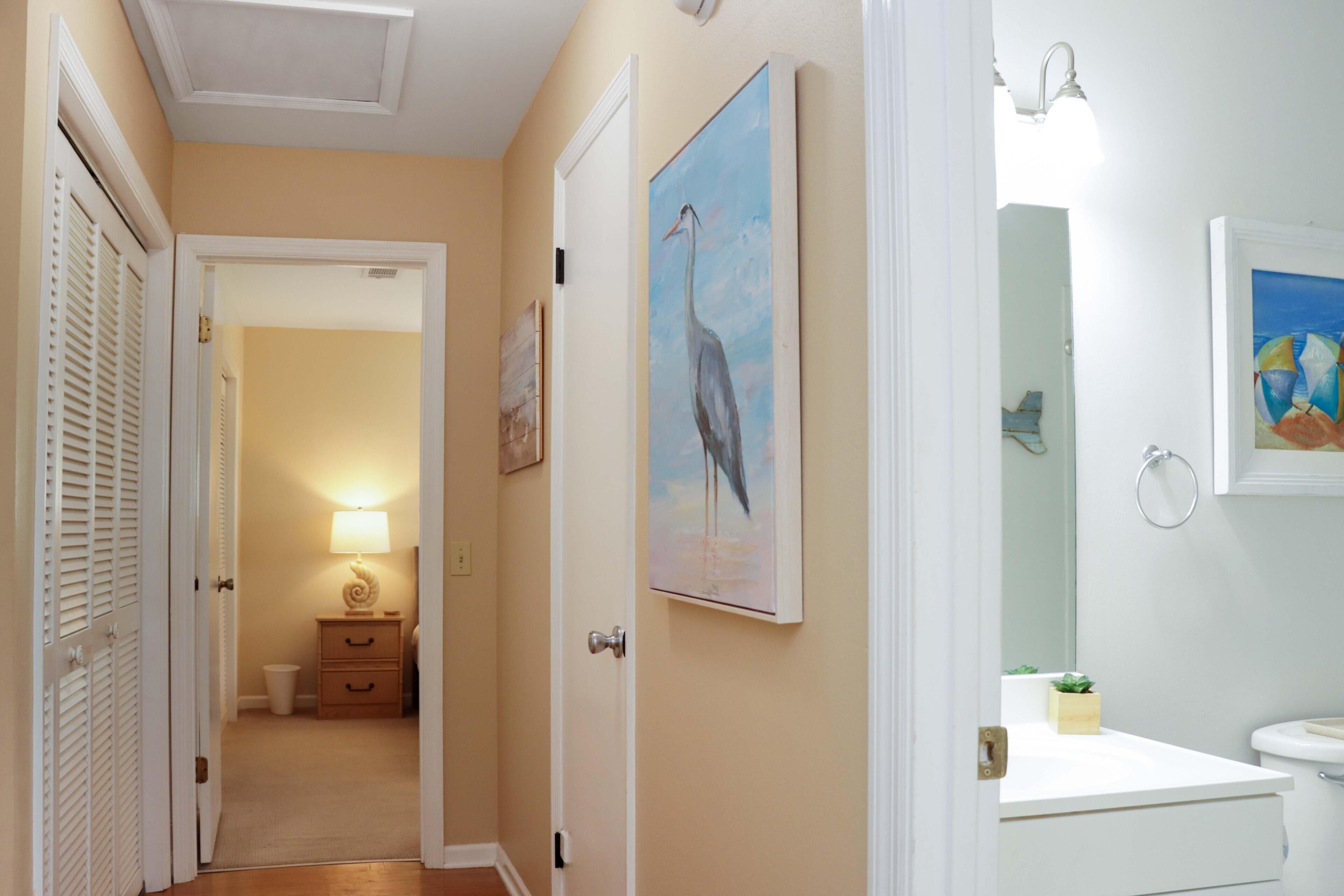A hallway with a door leading to a bedroom, a painting of a bird, a bathroom with a sink and mirror, and a nightstand with a lamp.