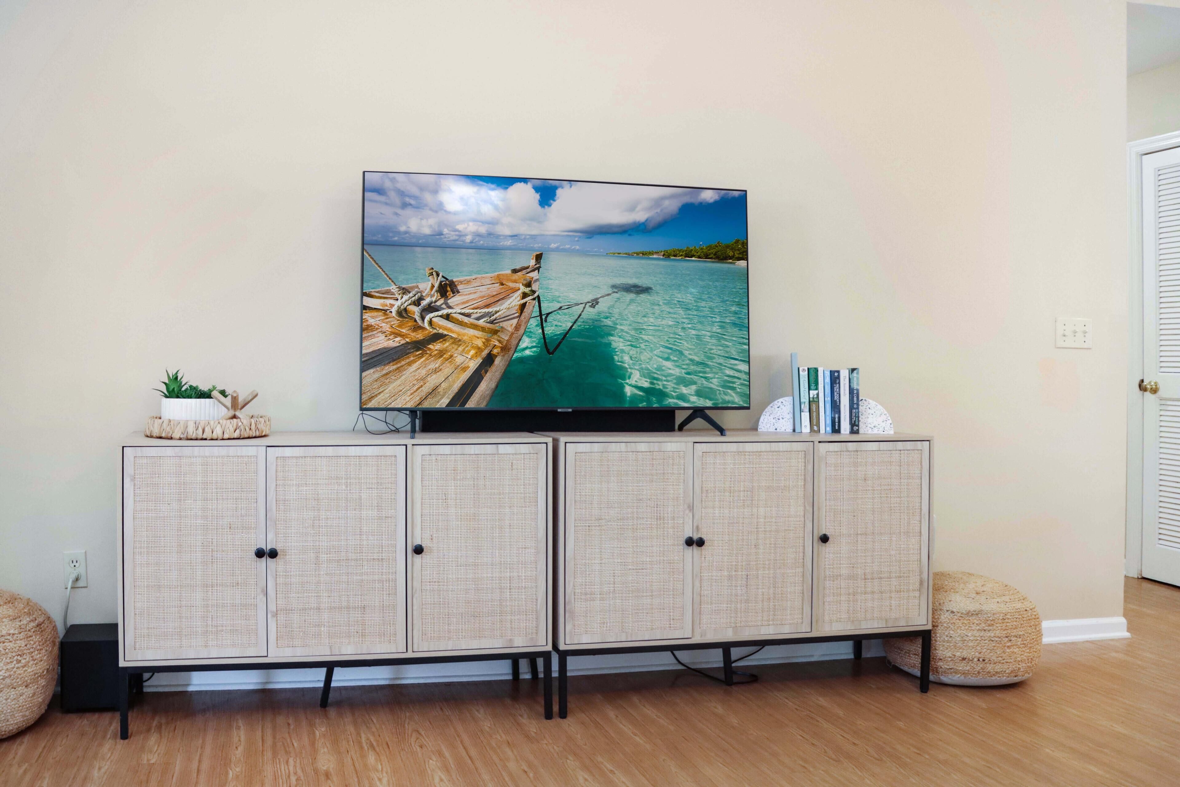 A flat-screen TV on a rattan cabinet with a tropical beach scene, a small plant, and books.