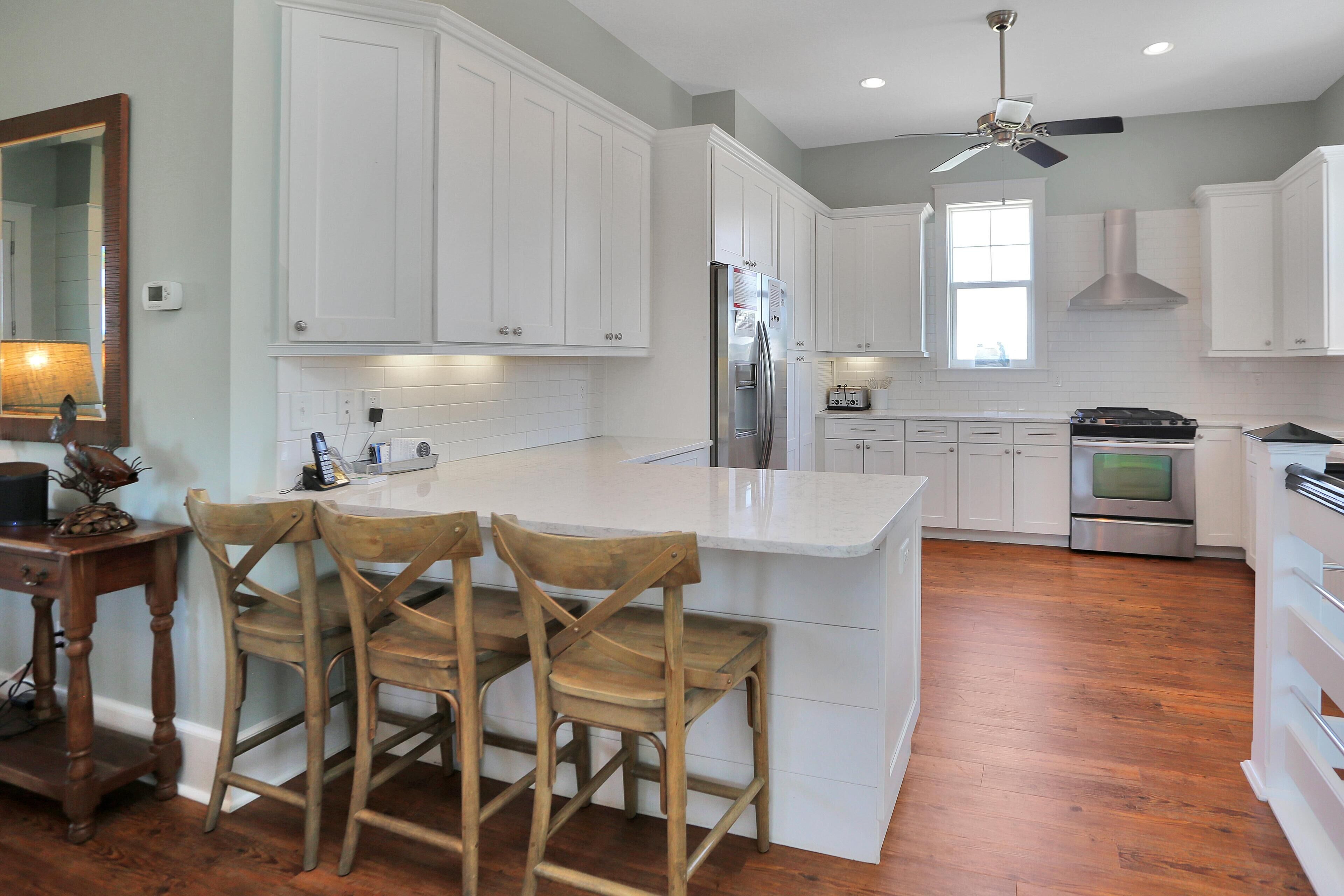 A kitchen with white cabinets, a large island with bar stools, a refrigerator, an oven, and a ceiling fan.
