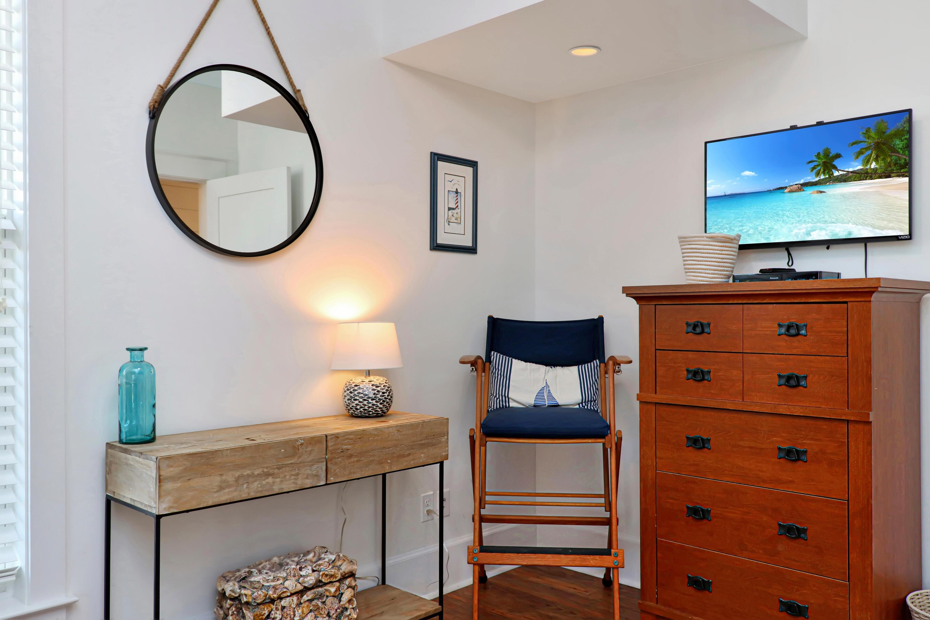 Une pièce comprenant une commode en bois, une chaise avec des coussins bleus, un miroir rond, un tableau encadré et un téléviseur diffusant une scène de plage.
