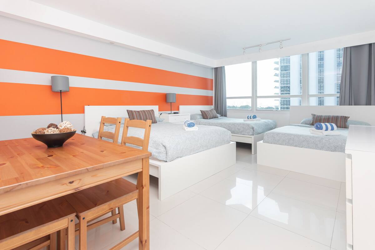 Quarto de hotel moderno com uma mesa de jantar de madeira, duas camas com roupa de cama branca, um abajur cinzento e vista da cidade através de grandes janelas.