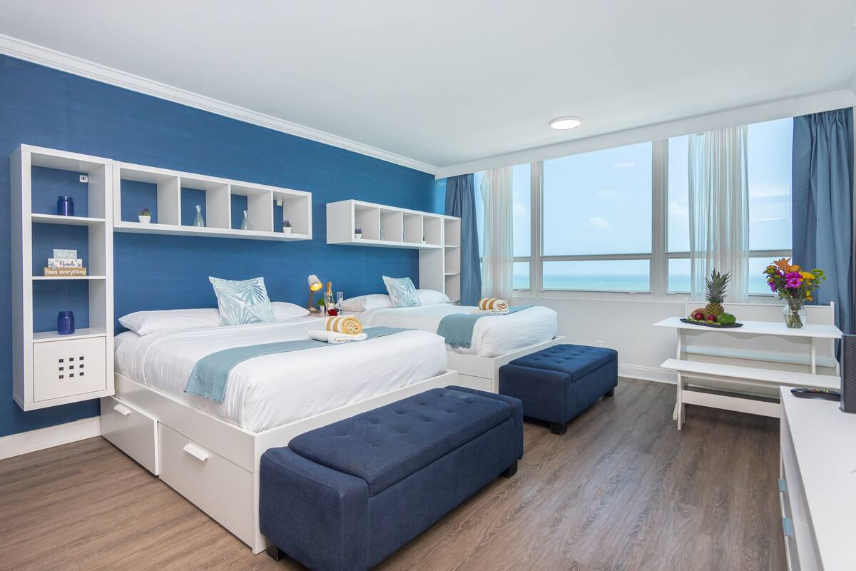 Quarto de hotel moderno com duas camas, uma janela grande com vista e uma mesinha com frutas.