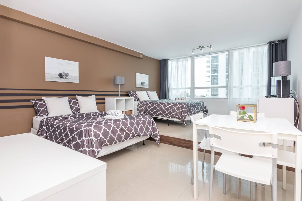 Quarto de hotel com duas camas, uma mesa de escritório e uma cadeira. Há duas mesas de cabeceira com luminárias.