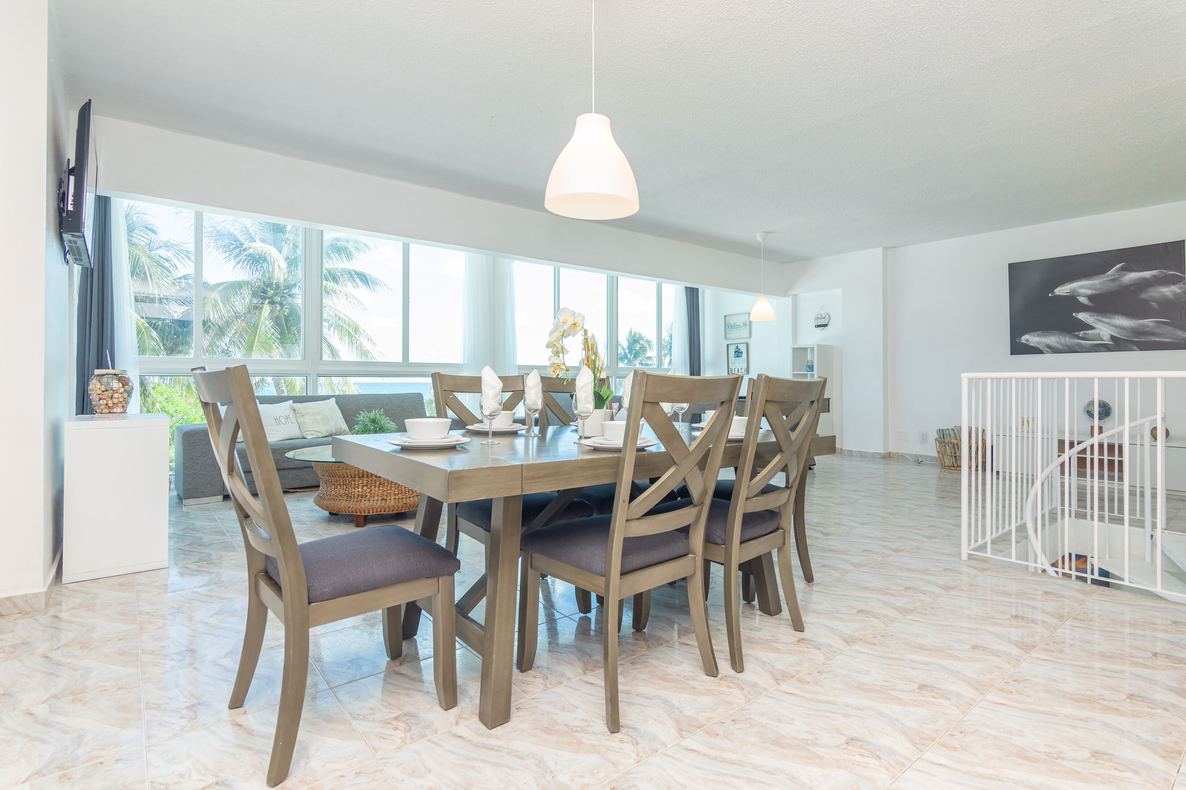 Un comedor con una mesa y sillas de madera, una lámpara colgante y ventanas amplias con vista a palmeras.