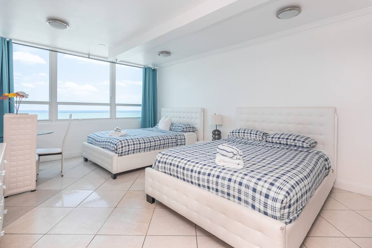 Quarto de hotel com duas camas, vista para o mar e uma grande janela com cortinas.