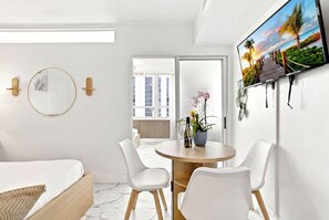 A modern hotel room with a bed, a round table with chairs, a TV on the wall, and a balcony with a view of buildings.