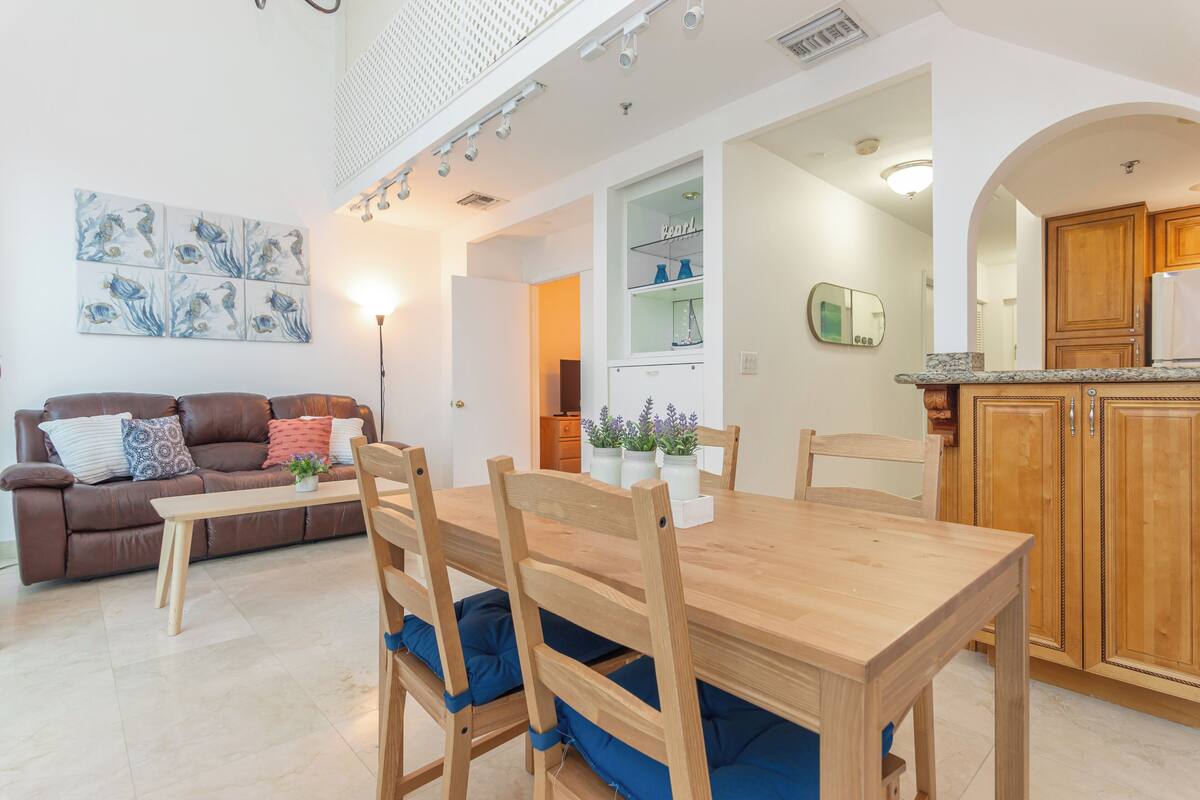 Sala de estar com sofá marrom, mesa de jantar de madeira e almofadas azuis. Há uma cozinha com armários de madeira e bancada.