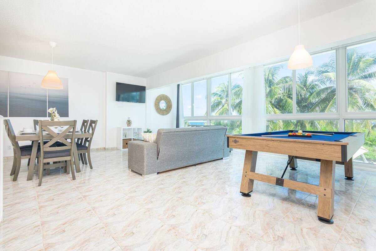 Uma sala de estar espaçosa com mesa de bilhar, um sofá cinzento e uma mesa de jantar com cadeiras.