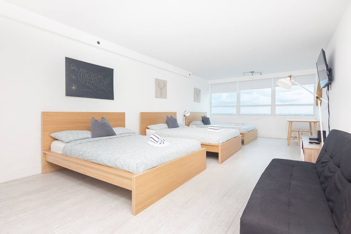 Quarto de hotel moderno com duas camas de madeira, um sofá preto, uma escrivaninha com luminária e uma janela grande que oferece uma vista.