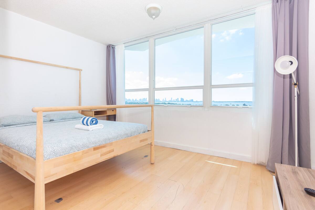Quarto minimalista com cama de madeira, uma janela ampla com vista para a cidade e uma mesa de cabeceira com abajur.