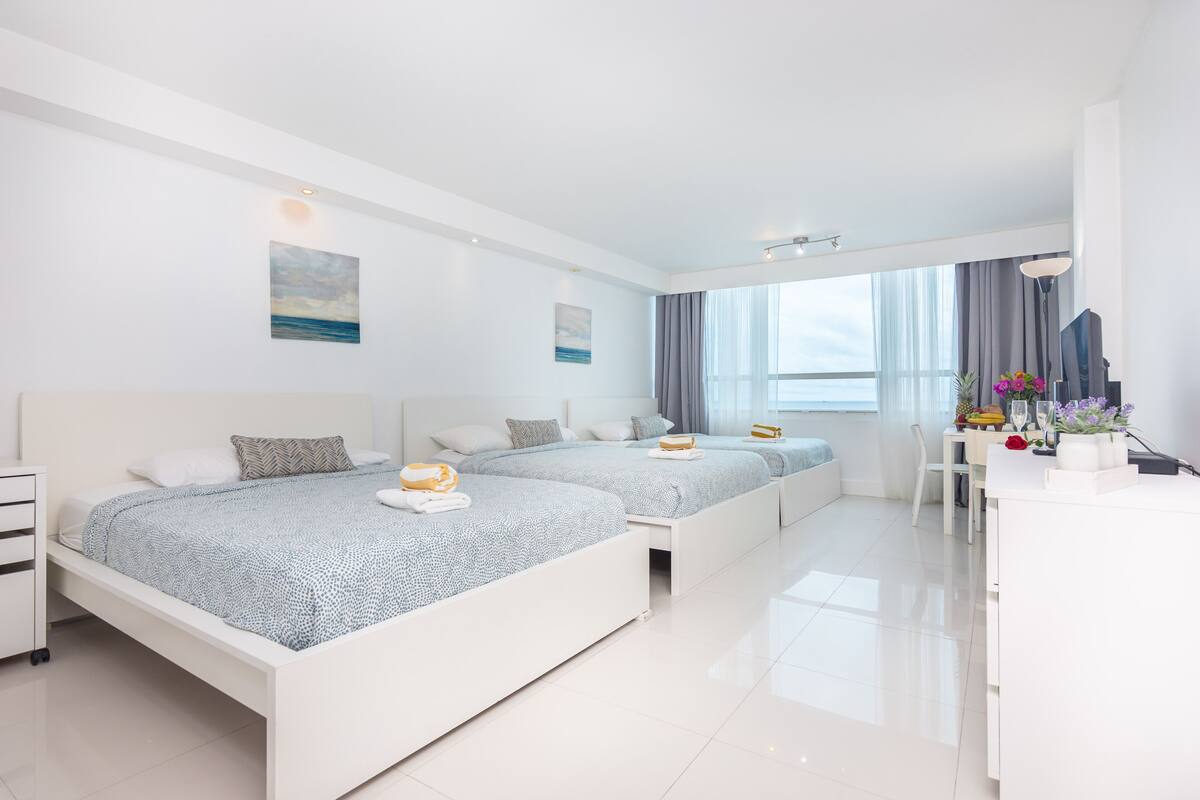 Quarto de hotel moderno com duas camas, uma mesa de trabalho e uma janela grande.