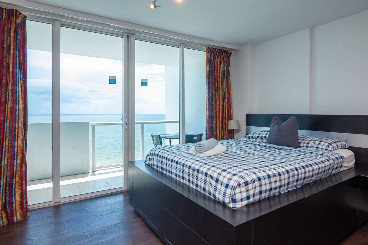 Quarto de hotel moderno com uma cama grande, vista para o mar e varanda com cortinas.
