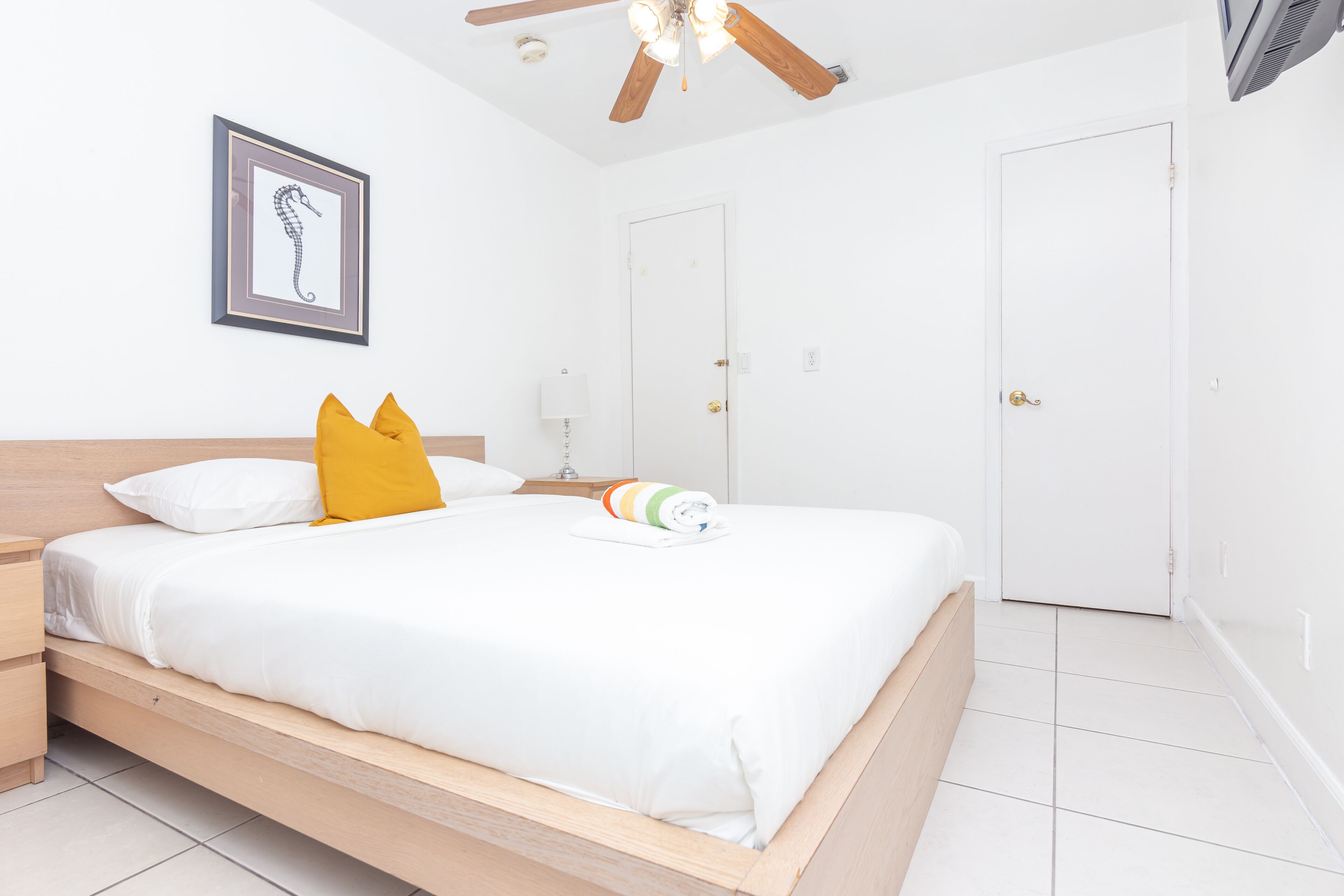 A neatly made bed with white bedding and a colorful towel, a wooden headboard, a framed seahorse artwork on the wall, and a ceiling fan.
