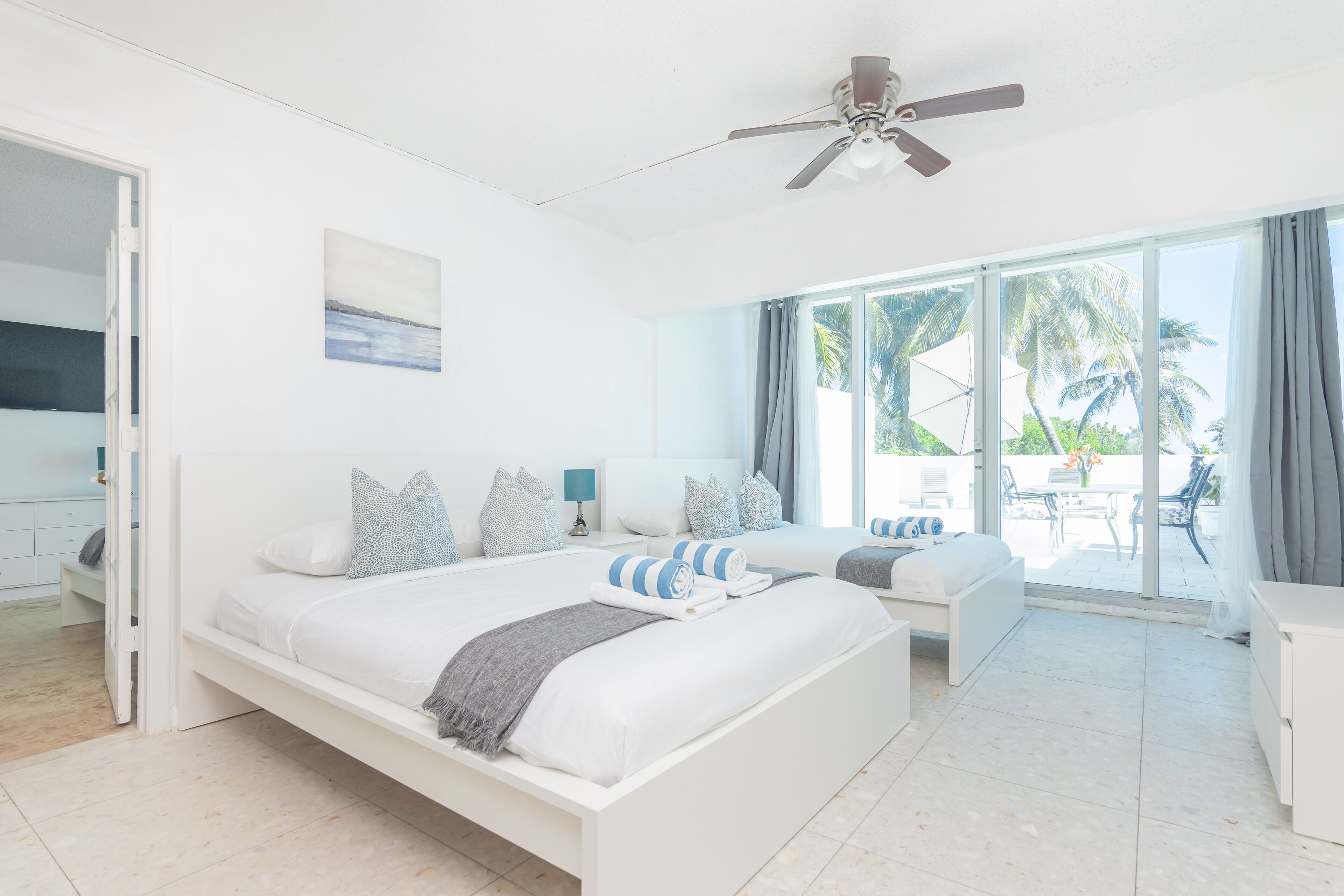 A modern hotel room with a large bed, a ceiling fan, a balcony with a view of palm trees, and a sliding glass door leading to a patio area.