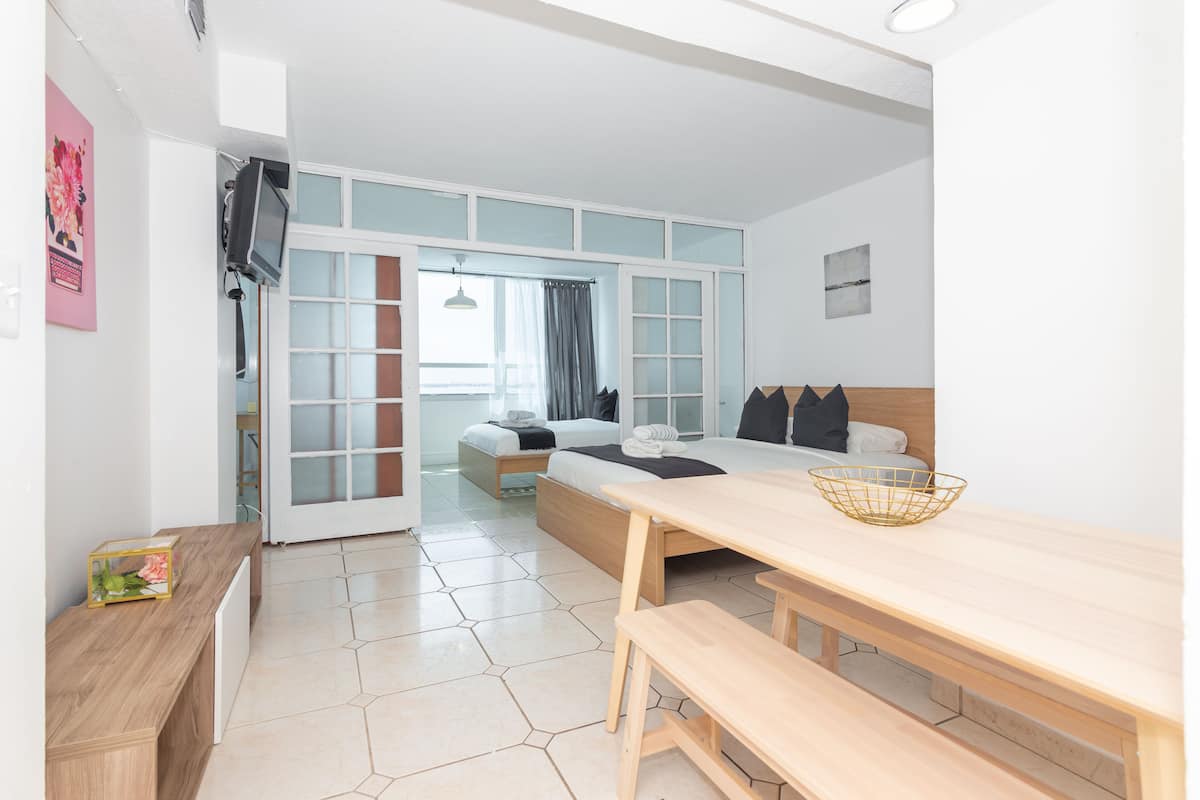 Quarto de hotel moderno com uma cama grande, um banco de madeira, uma mesa de jantar e uma TV fixada na parede.
