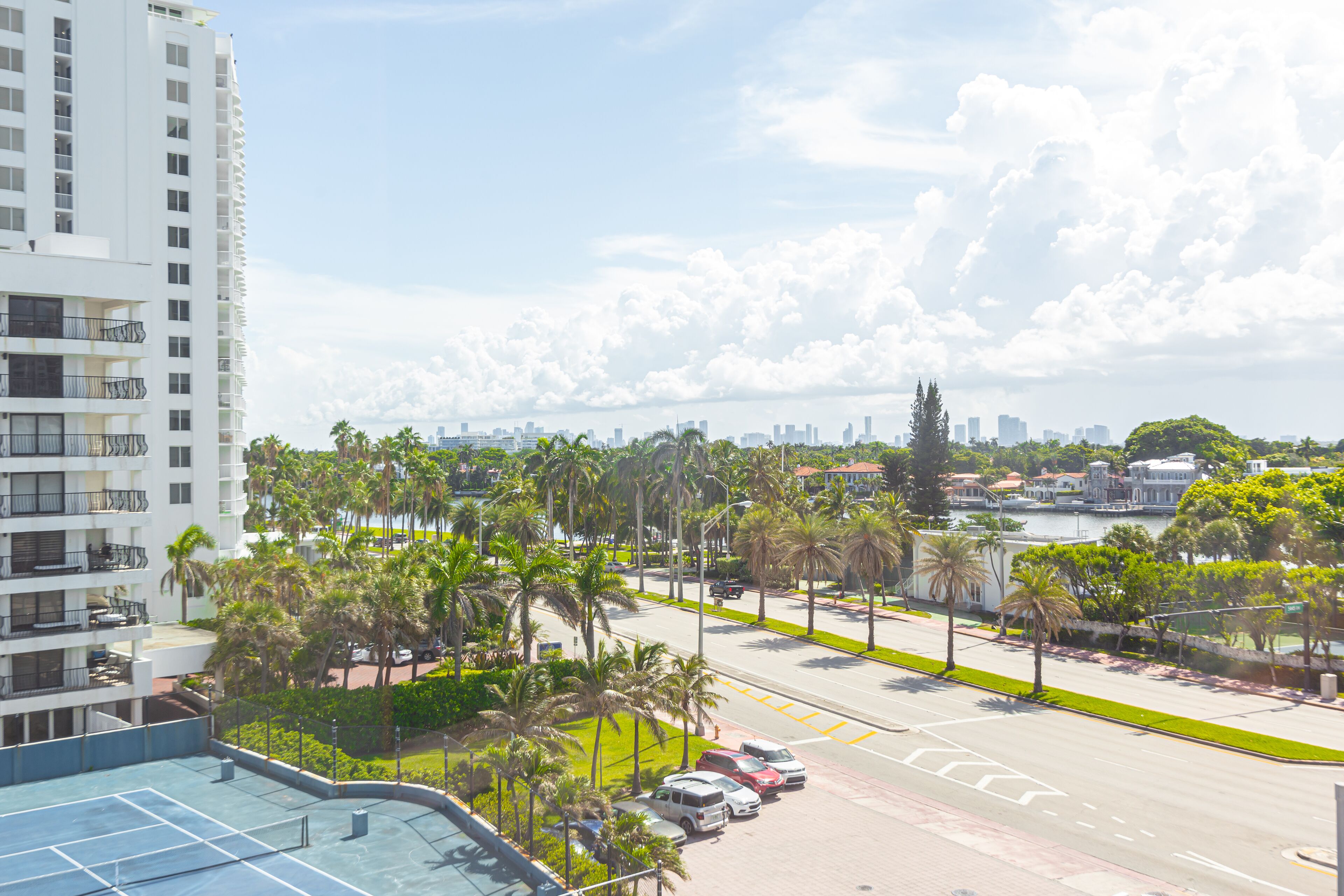 A high-rise building with multiple balconies, a clear view of a city skyline, and a street lined with palm trees.