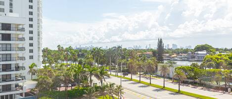 A high-rise building with multiple balconies, a clear view of a city skyline, and a street lined with palm trees.