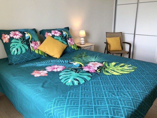 A bed with a tropical-themed bedspread, two pillows, a bedside lamp, and a chair.