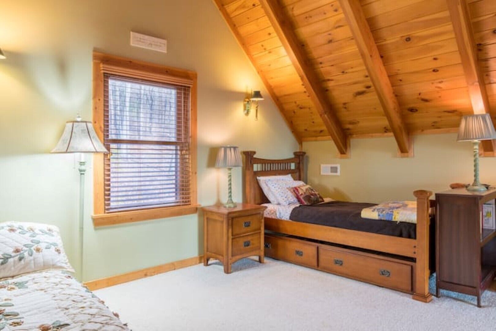 A bedroom with a wooden ceiling, a bed with a headboard, a nightstand, a lamp, a window with blinds, and a bedside table.