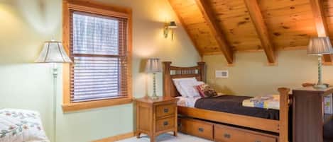 A bedroom with a wooden ceiling, a bed with a headboard, a nightstand, a lamp, a window with blinds, and a bedside table.