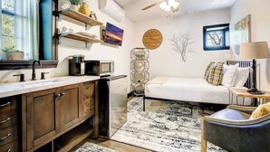 A compact bedroom with a bed, a desk, a chair, a ceiling fan, a clock, and a window. - Courtyard Studio at Bancroft Manor. Cozy Retreat! (Lusby)