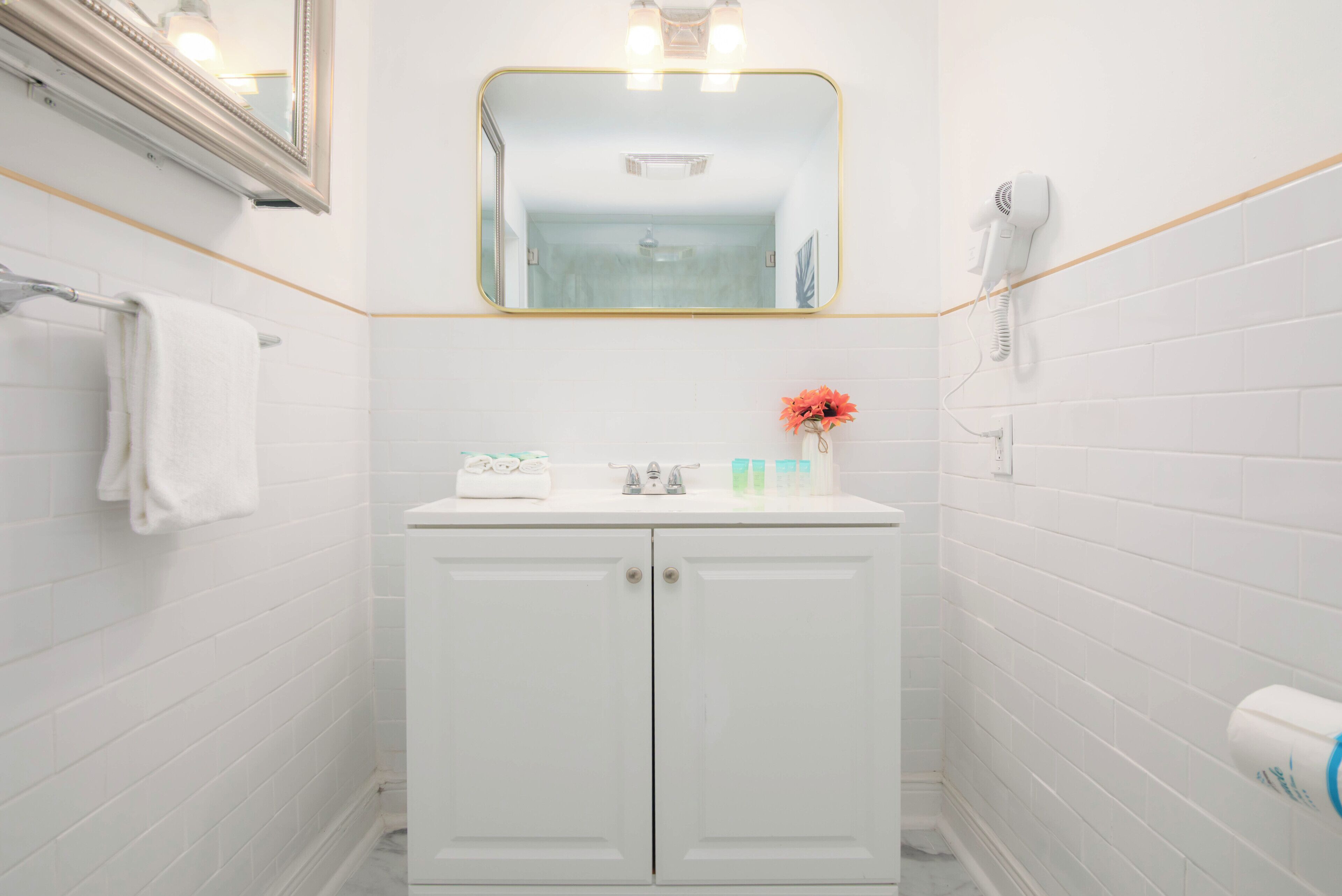 A bathroom with white tiled walls, a white vanity with a mirror above it, a towel rack, a hairdryer, and a small vase with flowers.