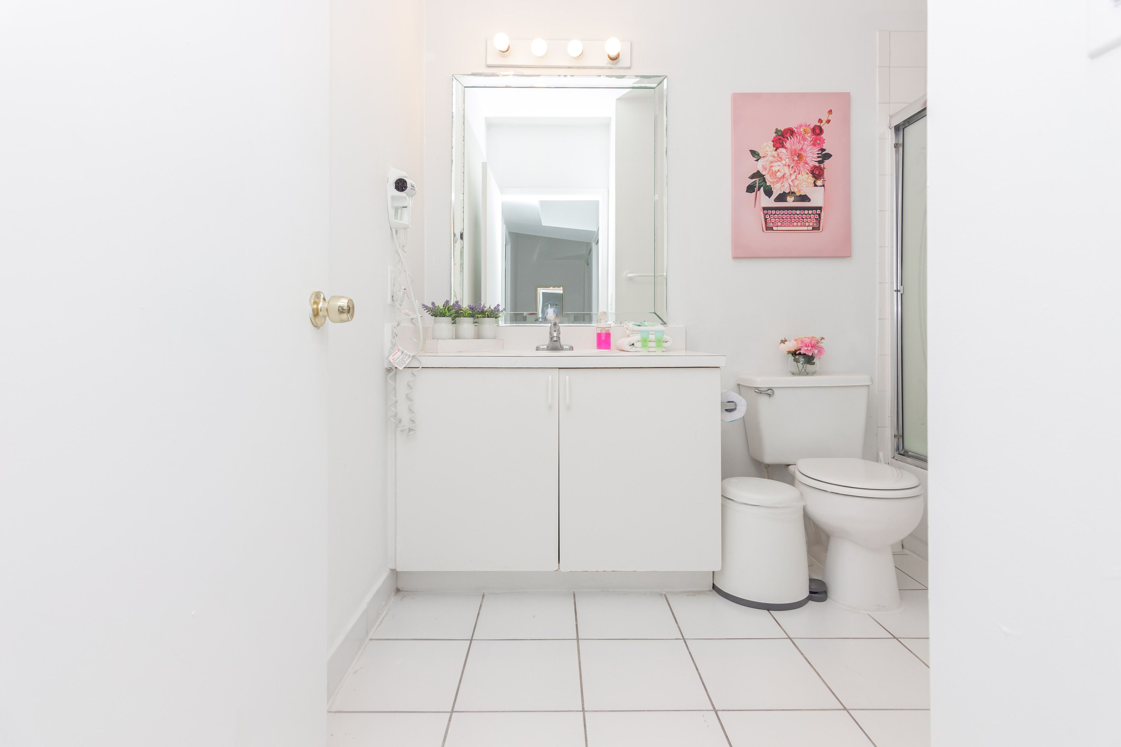 Un baño con lavamanos blanco, espejo y dos inodoros. Hay un cubo de basura y una lámpara en la pared sobre el espejo.