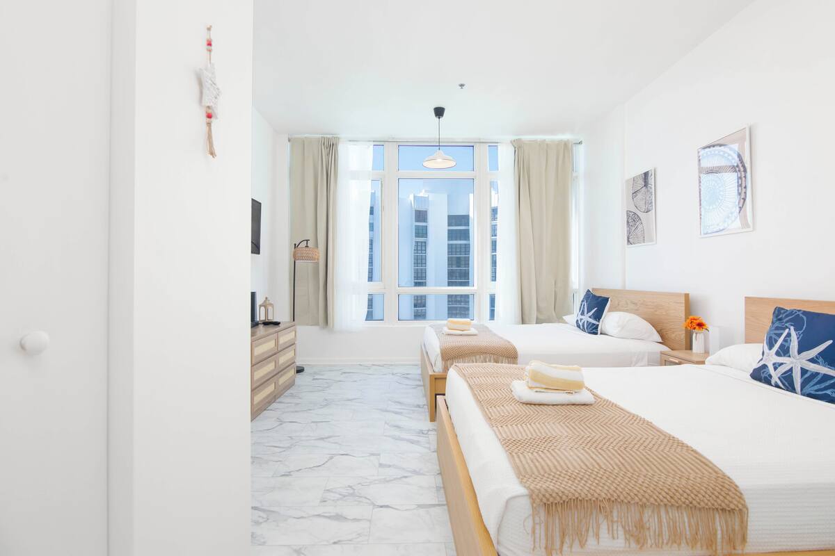 A hotel room with two beds, a large window, a wooden dresser, and artwork on the walls.