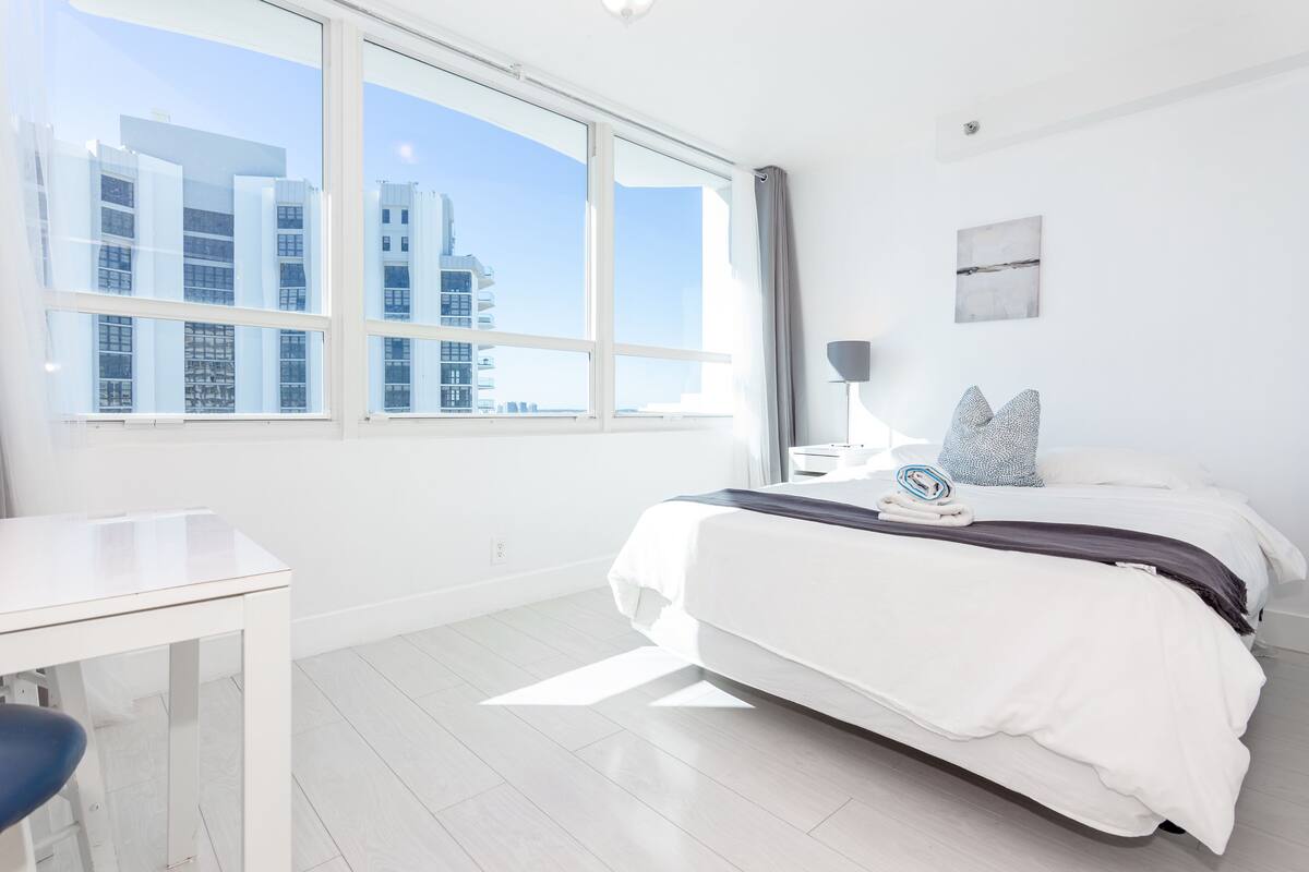 A modern hotel room with a large window, a neatly made bed, a small desk, and a lamp.
