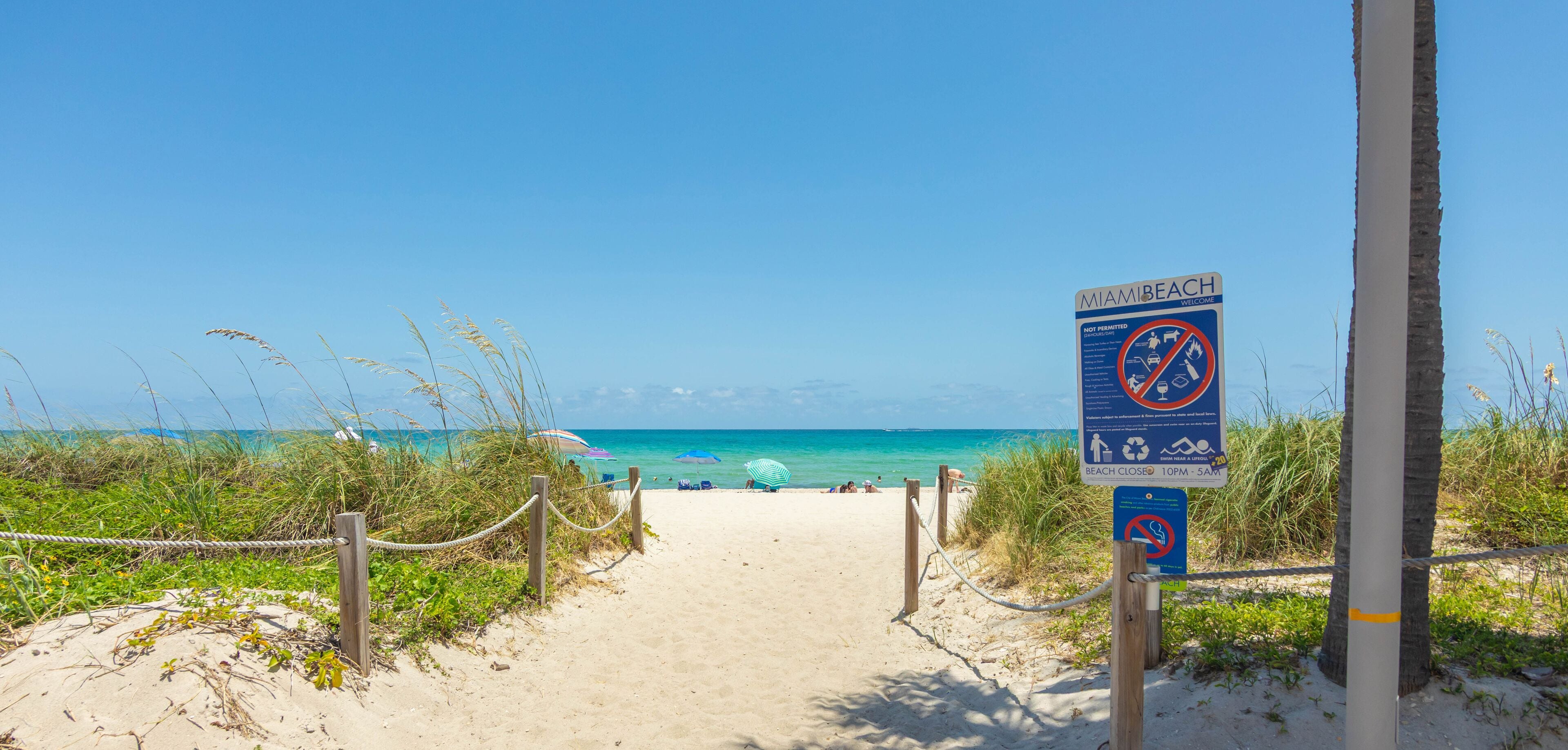 Ein Küstenpfad führt zum Meer, mit einem Schild, das die Schließzeiten des Strandes angibt.