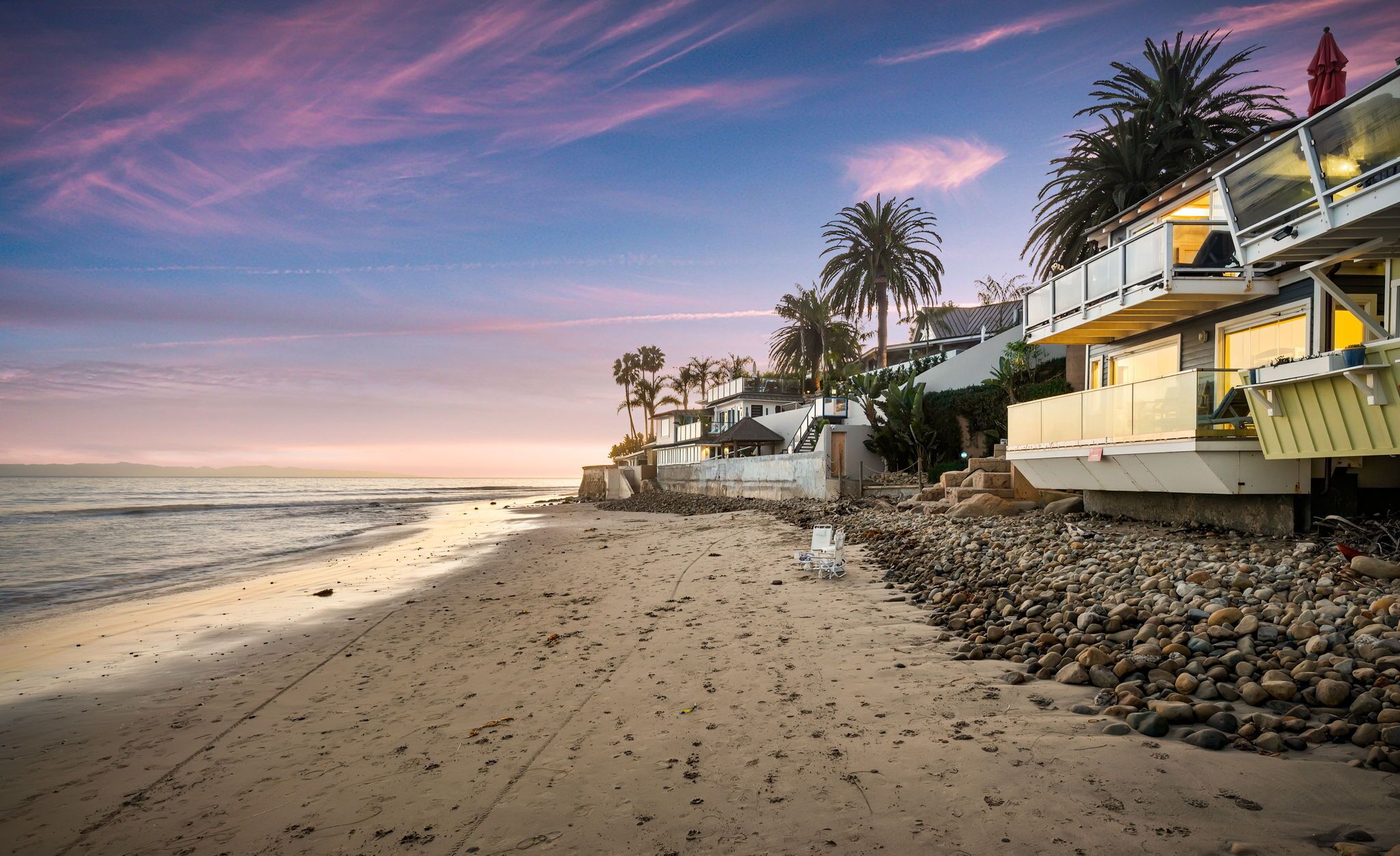 A beachfront property with a sandy beach, palm trees, and a modern building with a balcony overlooking the ocean.