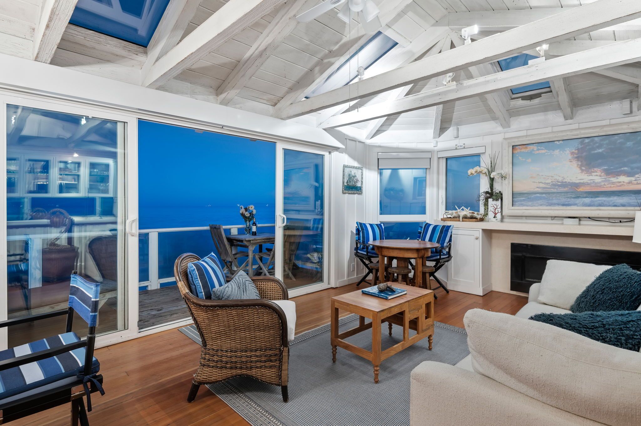 A spacious indoor area with a large sliding glass door leading to a balcony, wicker furniture, a wooden coffee table, and a painting of a beach scene.