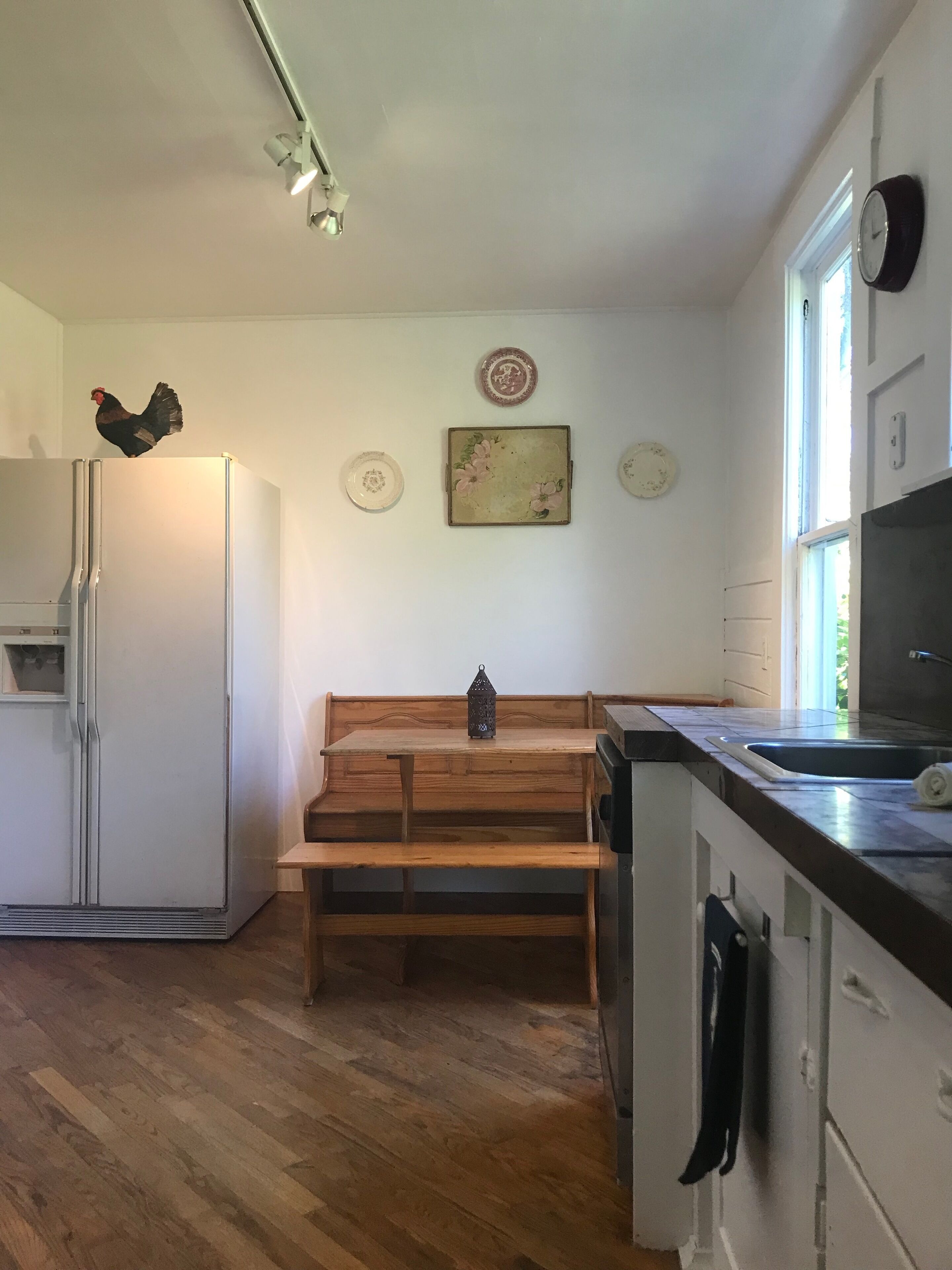 Una cocina con un banco de madera, un refrigerador, un lavamanos y adornos en la pared, incluyendo platos y un reloj.