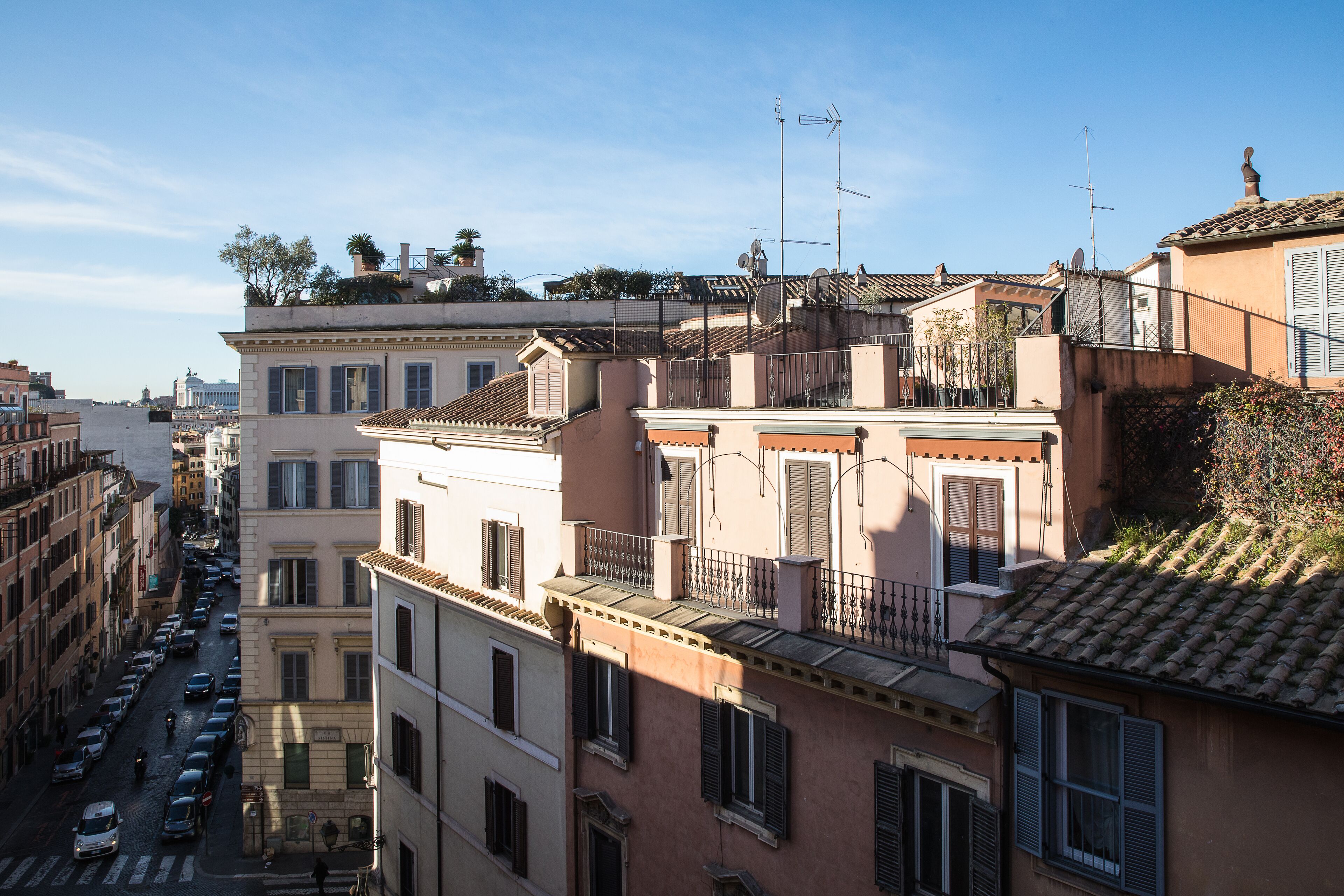 A row of traditional European buildings with balconies, a clear sky, and a street with parked cars.