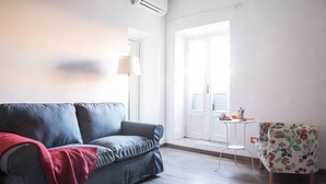 Quarto com sofá cinzento, manta vermelha, mesa pequena com abajur e cadeira estampada.