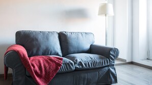 Um sofá cinzento com um cobertor vermelho, um abajur e uma janela com vista para o exterior.