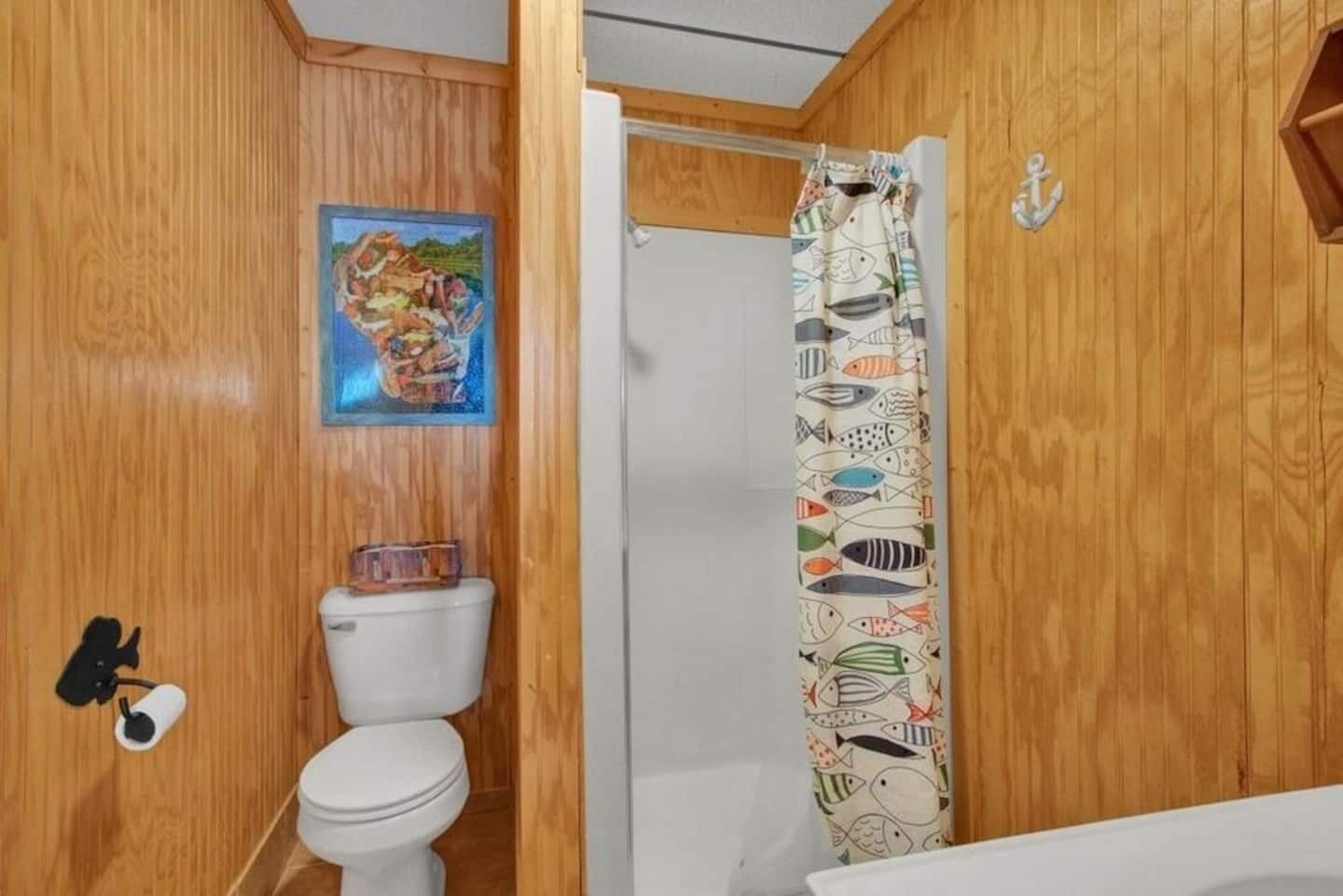 A wooden-paneled bathroom with a toilet, a shower curtain with fish patterns, and a small painting on the wall.