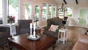 A spacious living room with a large wooden coffee table, a striped armchair, and a grey sofa. There are multiple windows providing natural light and a view of the outdoors. - Old Lake Charm with Million Dollar Views (Troutman)